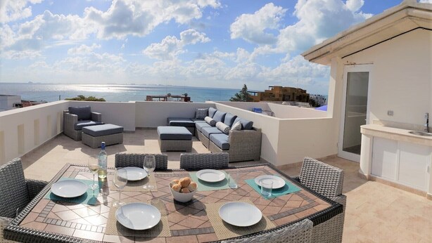 A rooftop terrace with outdoor seating, a dining table set with plates and glasses, and a view of the sea. - Experience Luxury on the Island of Isla Mujeres  (ISLA MUJERES)