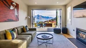 Una sala de estar moderna con sofá, una mesa de centro y chimenea. Hay un ventanal con vistas a montañas y una casa.