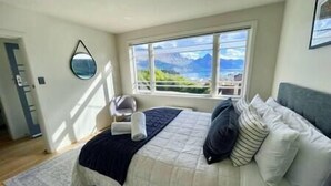 Un dormitorio con una cama grande, vistas de montañas a través de la ventana y una pequeña zona de estar con una silla y una mesa.