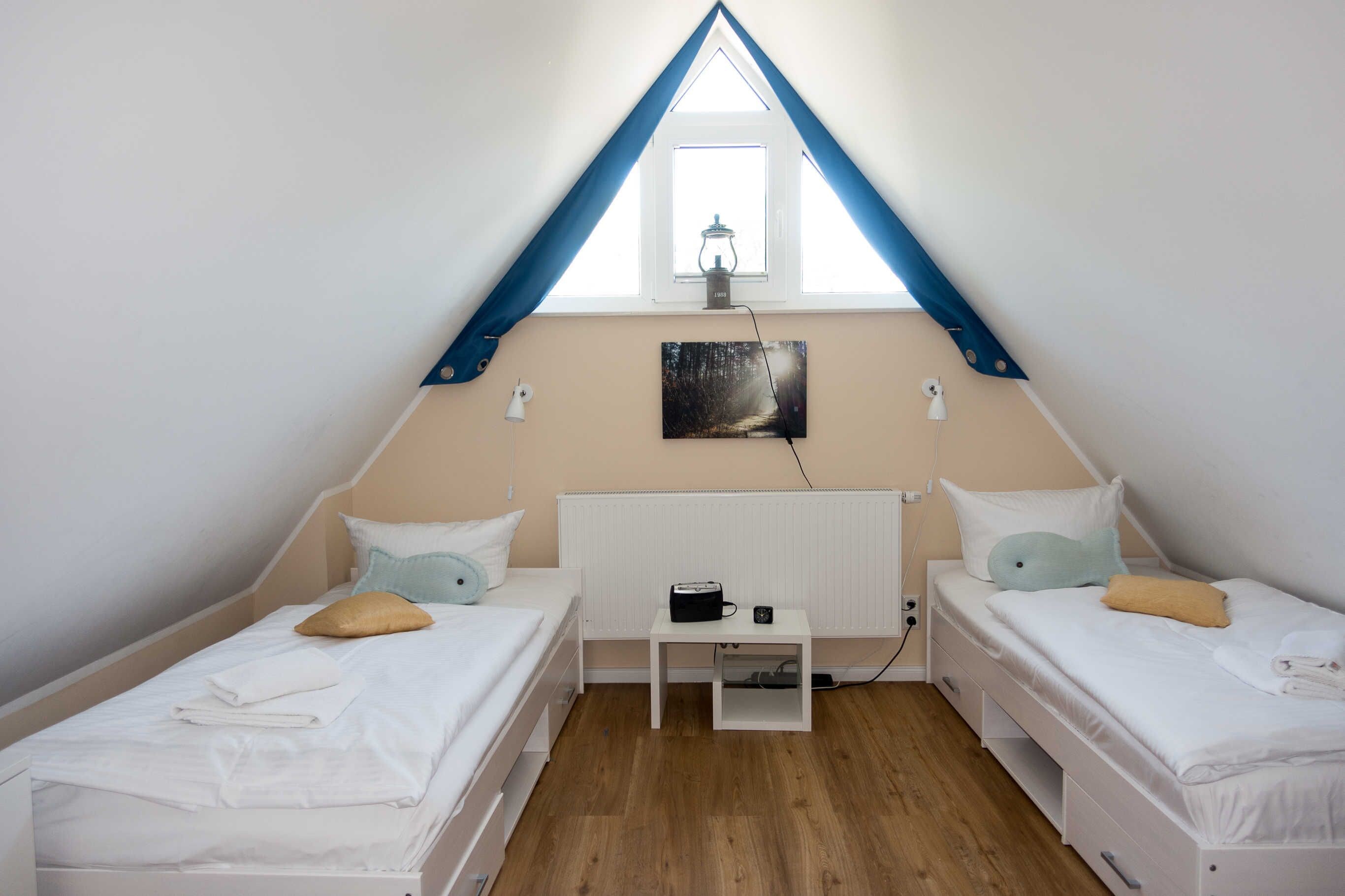 Una habitación pequeña y triangular con dos camas, un radiador, una mesa y una ventana.