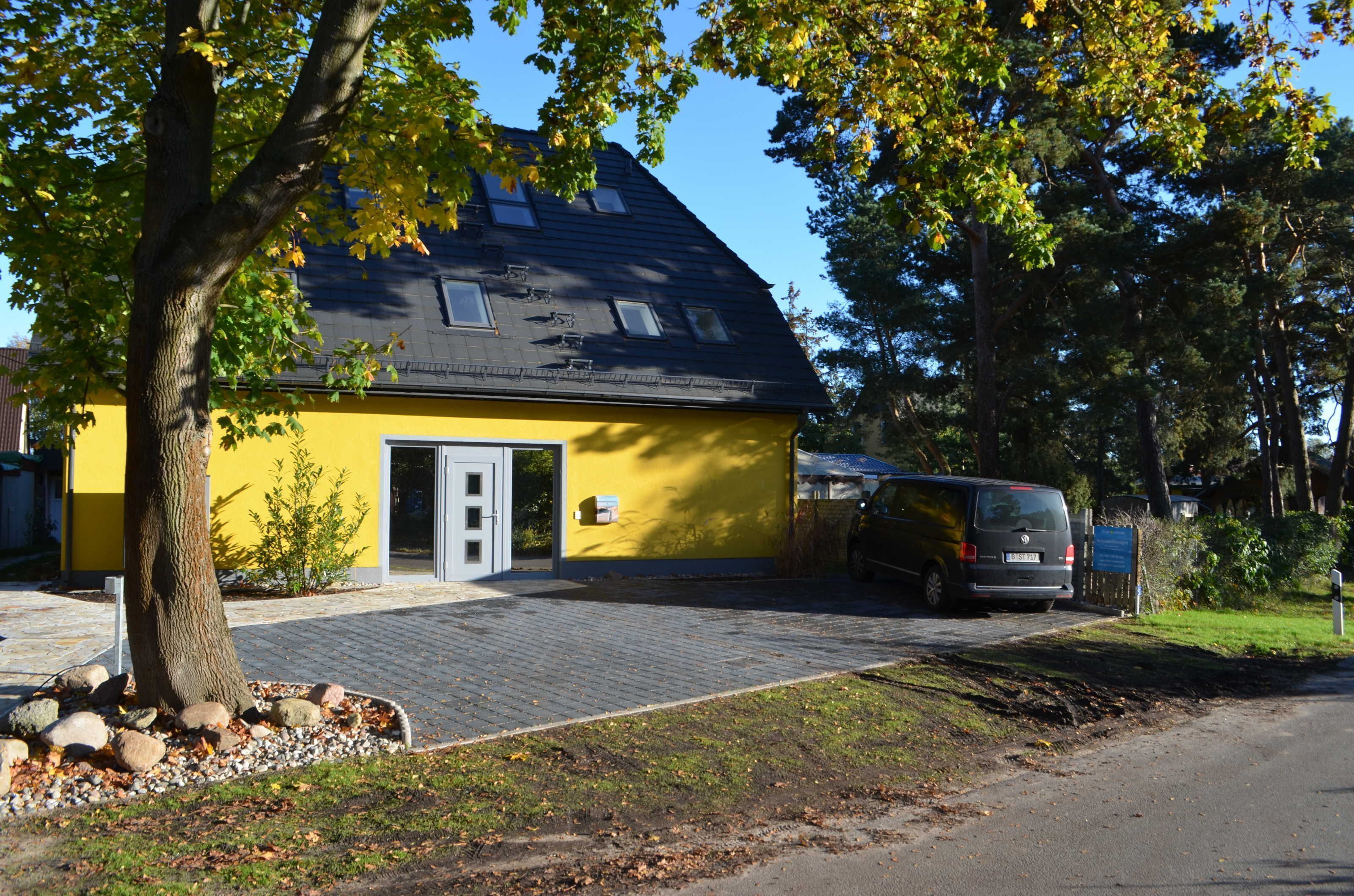 Un bâtiment jaune avec un toit sombre, un arbre au premier plan et un fourgon garé dans l’allée.