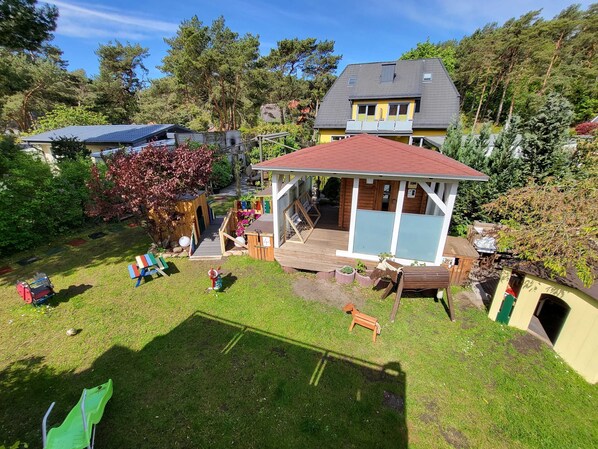 A residential property with a fenced backyard, a wooden playhouse, and a large house with a red roof.