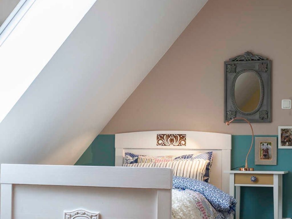 Ein Schlafzimmer mit schrägen Wänden, einem Bett mit gemusterten Betttextilien, einem Nachttisch mit Lampe und einem wandmontierten Spiegel.