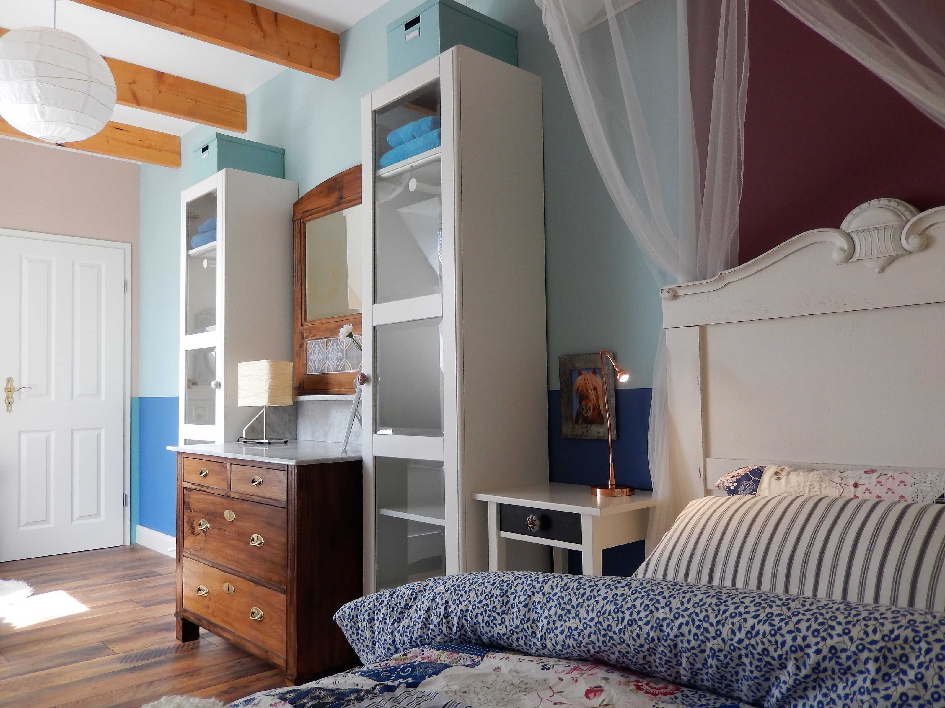 A bedroom with a wooden dresser, a bed with patterned bedding, a nightstand, a lamp, and a wardrobe.
