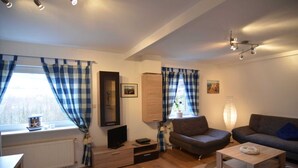 Ein modernes Wohnzimmer mit einer Couch, Sesseln, einem Couchtisch und einem Fernseher. Vorhanden sind Vorhänge, eine Lampe und eine Heizung.