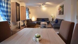Ein Wohnzimmer mit einer Couch, einem Couchtisch und einem Esstisch mit Tischdekoration. Eine Lampe und ein Gemälde befinden sich an der Wand.