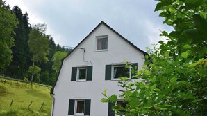 Ein weißes Haus mit grünen Fensterläden, eine kleine Veranda mit Tisch und Stühlen und Blick auf einen grünen Hügel.