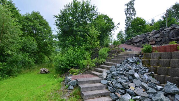 Eine steile Steintreppe führt einen grasbewachsenen Hang hinauf, umgeben von dichtem Grün und einer Steinmauer.