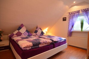 Un dormitorio con cama, ropa de cama morada, una mesita de noche, una lámpara, una ventana con cortinas moradas y un radiador.