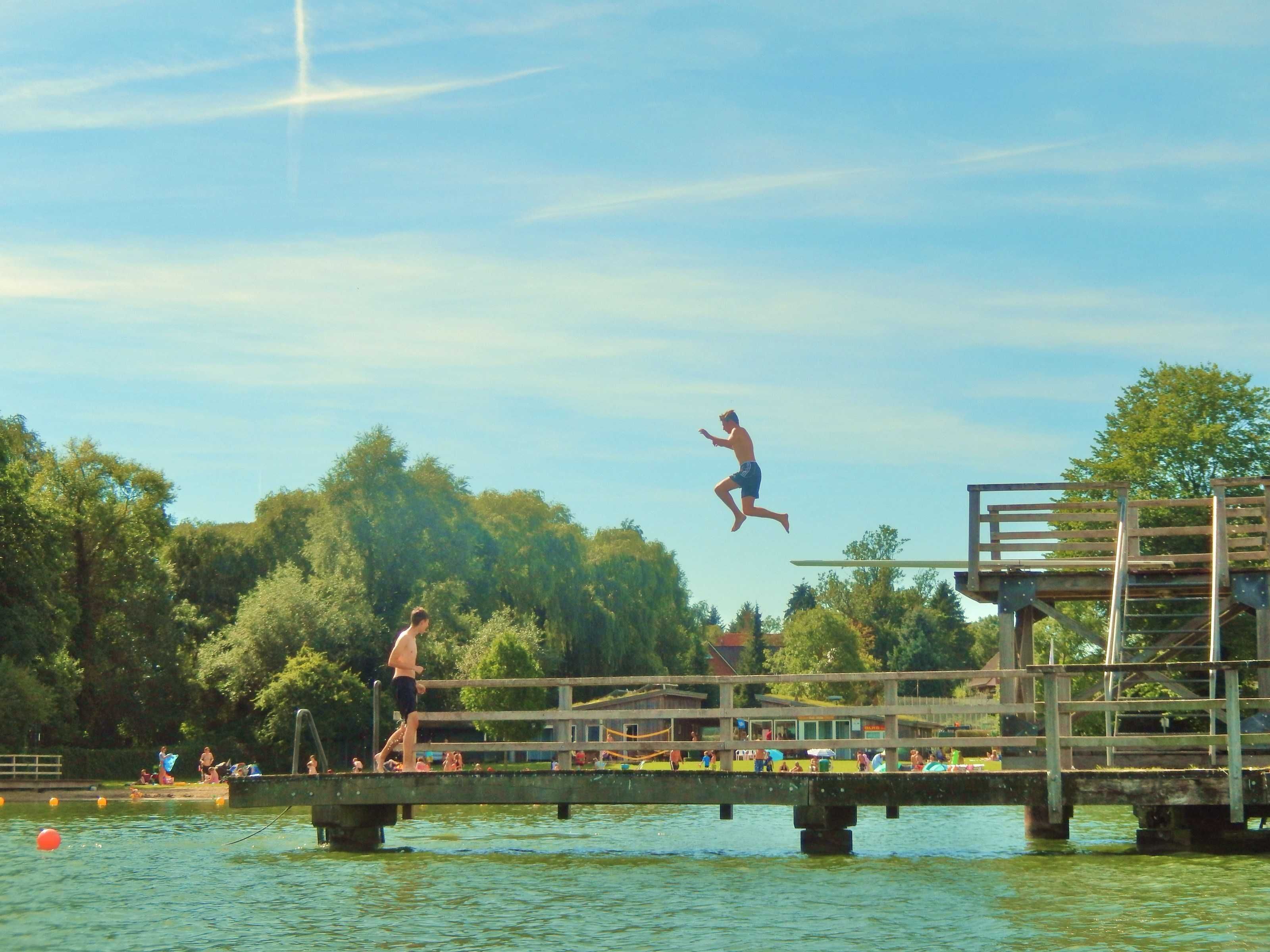 A wooden dock extending over water with people on it, a diving board, and a clear sky.