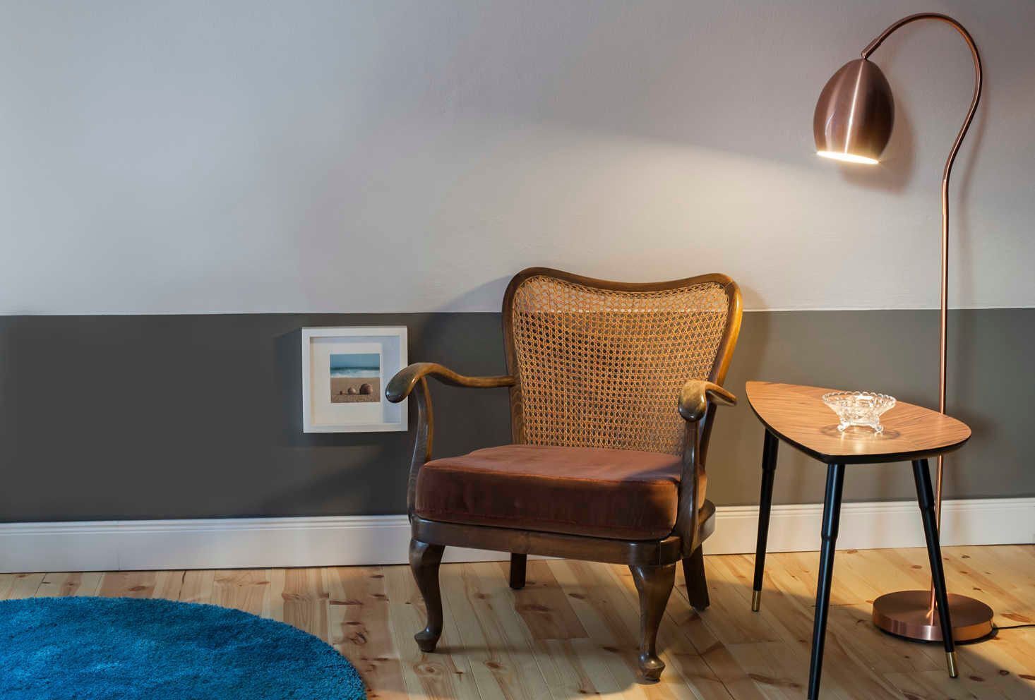 A room with a wooden chair, a small table with a glass bowl, a floor lamp, and a framed picture on the wall.