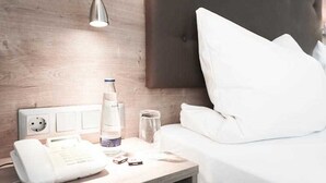 A hotel room with a wooden headboard, a bedside table with a lamp, a phone, a bottle of water, and a glass jar. - Double room - Hotel Poinger Hof (Poing)