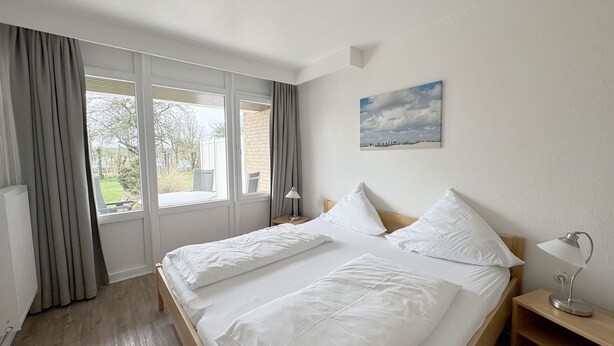 A hotel room with a large bed, two bedside tables, a window with curtains, and a painting on the wall. - Luna22 (room category) - Luna III (Sankt Peter-Ording)