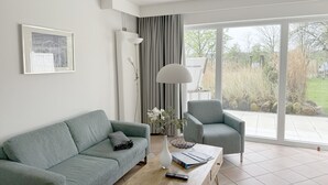 A modern living room with a grey sofa, a matching armchair, a coffee table, and a large window with a view of a garden. - Luna22 (room category) - Luna III (Sankt Peter-Ording)