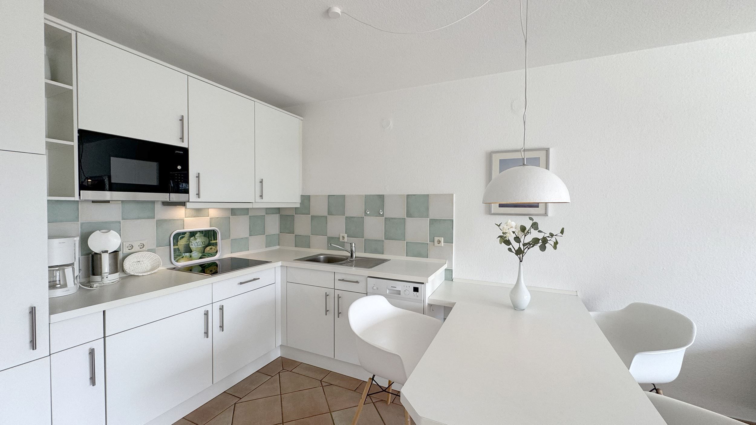 Una cocina moderna con gabinetes blancos, un microondas integrado y un lavabo. Hay una mesa de comedor blanca con dos sillas y un florero con flores.