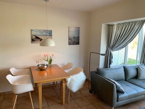 Ein Essbereich mit Holztisch, weißen Stühlen, einer Schüssel und einem Blumenstrauß. Im Wohnbereich befindet sich ein graues Sofa und eine Stehlampe.