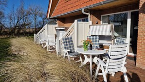 An outdoor seating area with white and blue checkered chairs, a round table with a vase of flowers, and a bottle of wine. - Luna IV - Luna Apartment 30 (Sankt Peter-Ording)