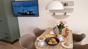 A dining area with a table set for breakfast, including bread, eggs, and beverages. A television displaying a ship is mounted on the wall. - Luna IV - Luna Apartment 30 (Sankt Peter-Ording)