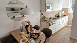 A modern kitchen with white cabinets, a microwave, and a sink. There is a dining table with a vase of flowers and a bottle of milk. - Luna IV - Luna Apartment 30 (Sankt Peter-Ording)