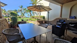 Un área de patio al aire libre con mesa y sillas, un paraguas grande y vistas a un jardín ajardinado con palmeras.