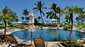Un área junto a la piscina con sillones, sombrillas y palmeras.