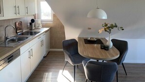 A modern kitchen with a dining area, featuring a wooden table, grey chairs, a white pendant light, and a vase with flowers. - Luna IV - Luna apartment 36 (Sankt Peter-Ording)