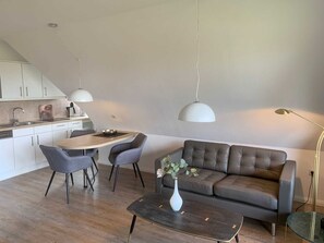 Una cocina moderna con armarios blancos, un comedor con una mesa de madera y dos sillas, un sofá gris y una pequeña mesa de centro con un jarrón.