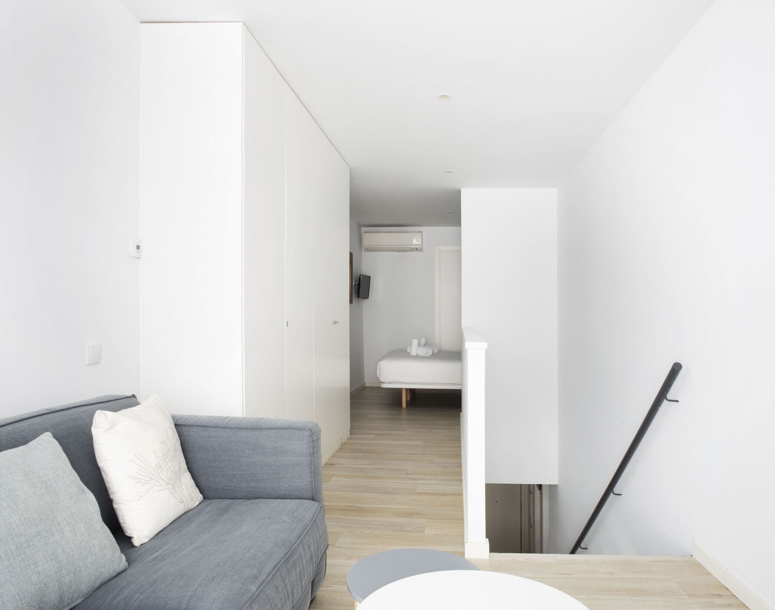 Un salon moderne et épuré, avec un canapé gris, un fauteuil blanc et une petite table ronde. Un escalier mène à une salle de bain dotée d’un lavabo blanc et d’un porte-serviettes.