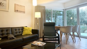 A modern living room with a leather sofa, a glass coffee table, a dining area with a wooden table and chairs, and large windows offering a view of greenery. - Luna20 (room category) - Luna II (Sankt Peter-Ording)