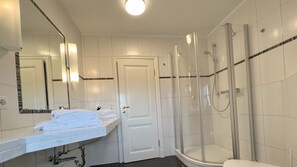 A bathroom with a white tiled wall, a glass-enclosed shower, a white sink with a mirror, and a white door. - Maren (Room category) - Maren (Sankt Peter-Ording)