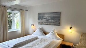 A neatly made bed with white linens, a painting of a landscape on the wall, and two wall-mounted lamps. - Farmhouse - Farmhouse apartment 25 (Sankt Peter-Ording)