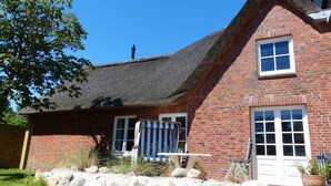 A traditional brick house with a steeply pitched thatched roof, a small garden with rocks and plants, and a white door. - Farmhouse - Farmhouse apartment 25 (Sankt Peter-Ording)
