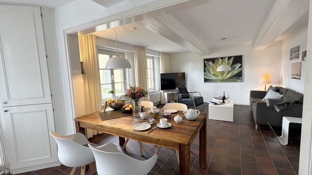A modern living room with a wooden dining table, white chairs, a grey sofa, and a TV. - Farmhouse - Farmhouse apartment 25 (Sankt Peter-Ording)