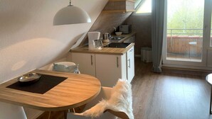 A compact kitchen with a dining area, a small refrigerator, a coffee maker, and a window with a view of a wooden deck. - Luna IV - Luna Apartment 33 (Sankt Peter-Ording)