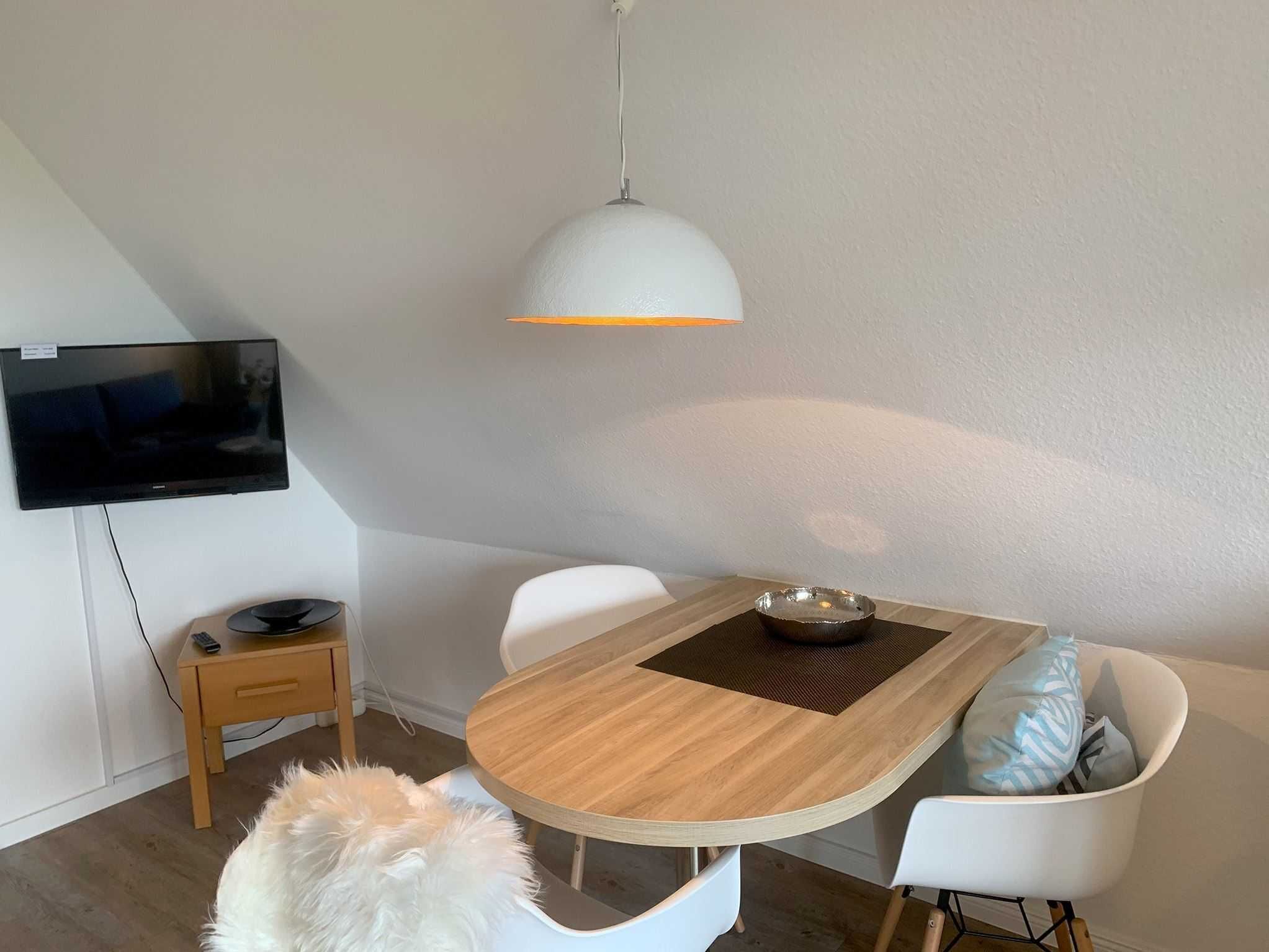 A modern dining area with a wooden table, white chairs, a television mounted on the wall, and a white pendant light.