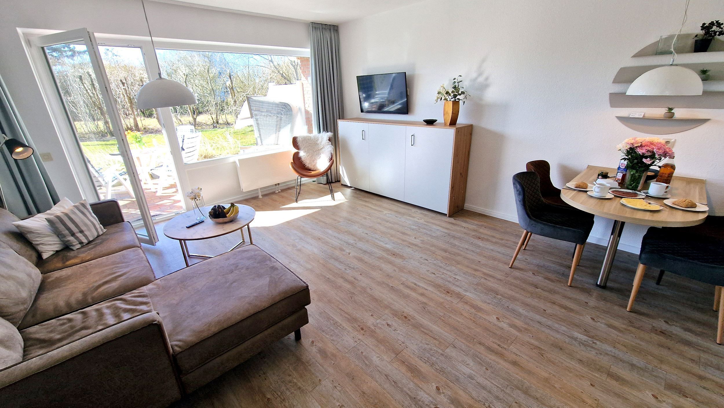 A modern living room with a sofa, a dining table with chairs, a TV, and a large window with a view of trees.