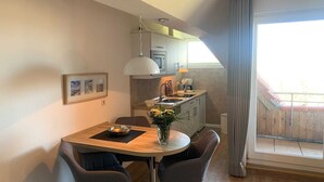 A dining area with a round table, two chairs, a vase with flowers, and a kitchen in the background. - Luna IV - Luna apartment 35 (Sankt Peter-Ording)