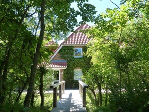 A wooden pathway leading to a house with a tiled roof, surrounded by lush greenery.