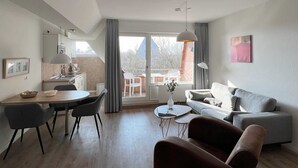 A modern living room with a grey sofa, a dining table with chairs, and a kitchen area in the background. - Luna IV - Luna apartment 35 (Sankt Peter-Ording)