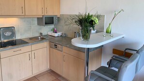 A compact kitchen with wooden cabinets, a microwave, a sink, and a small dining area with a chair. - Luna12 (Room category) - Luna I (Sankt Peter-Ording)