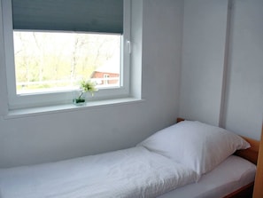 Une petite chambre avec un lit simple, une fenêtre et une petite table sur laquelle est posé un vase de fleurs.