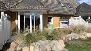 A house with a sloped roof, a front porch with chairs, and a small garden with rocks and grass. - Luna12 (Room category) - Luna I (Sankt Peter-Ording)