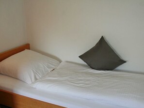 Ein Einzelbett mit Holz-Kopfstück und zwei Kissen, eines weiß, eines dunkelgrau, vor einer schlichten Wand.