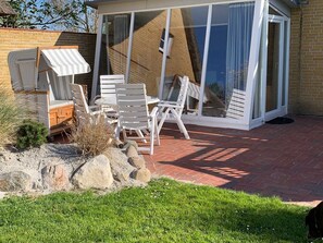 Eine Terrasse mit weißen Stühlen und einem Tisch, ein kleiner Gartenbereich mit Steinen und Gras sowie eine Glastür zu einem überdachten Außenbereich.