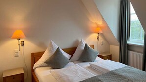 Um quarto com teto inclinado, duas luminárias de cabeceira, uma cama com roupa de cama branca e almofadas estampadas, e uma janela com cortinas.