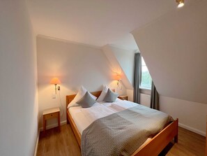 Quarto com teto inclinado, cama com almofadas, duas luminárias de cabeceira e janela com cortinas.