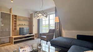 Uma sala de estar moderna com televisão, mesa de centro de vidro, sofá, poltrona e luminária pendente.