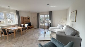 A living room with a sofa, a glass coffee table, a TV, a dining table with chairs, and a bookshelf. - AH1a (room category) - Ahndole Hollken (Sankt Peter-Ording)