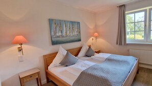 A hotel room with a bed, two bedside lamps, a painting, a window, and a radiator. - AH1a (room category) - Ahndole Hollken (Sankt Peter-Ording)