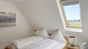 A bedroom with a bed, pillows, a nightstand, a lamp, a window, and a painting on the wall. - Luna19 (Room category) - Luna II (Sankt Peter-Ording)